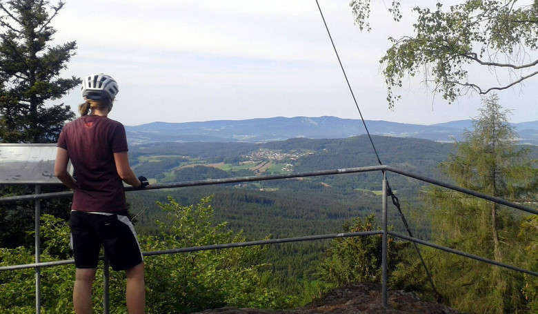 Frau mit Helm sieht auf einem Aussichtspunkt über das ARBERLAND hinab